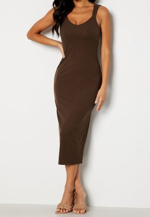 Frau trägt ein ärmelloses, geripptes, braunes Midikleid mit V-Ausschnitt, posiert mit einer Hand in der Hüfte und trägt geflochtene, hautfarbene Absätze.