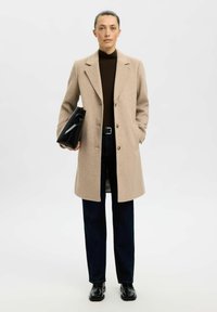 Beige wollen jas met grote revers, twee knopen en zijzakken, gedragen over een bruine coltrui en donkere spijkerbroeken, met een zwarte tas.