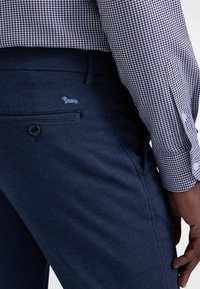 I pantaloni in denim blu presentano una tasca posteriore con un piccolo ricamo di un dachshund azzurro e sono abbinati a una camicia a quadri con motivo.