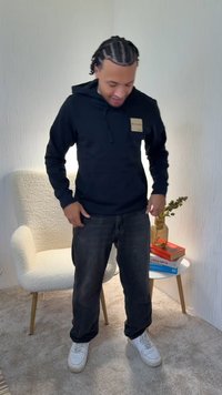 Černá mikina s kapucí s přední kapsou a štítkem, doplněná tmavými džínami a bílými teniskami. Pozadí zahrnuje krémově zbarvené křeslo a knihy.