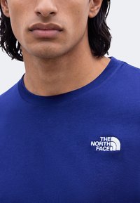 Gros plan d'une personne portant un t-shirt bleu avec un logo blanc "The North Face" sur la poitrine.