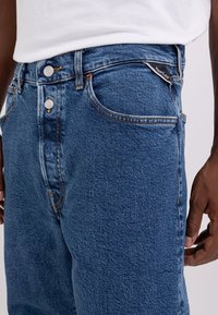 Jeans de mezclilla azul con corte recto, que cuentan con dos botones plateados y bolsillos cosidos. La tela tiene un acabado texturizado.