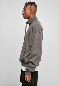 Grå høyhalset sweatshirt med lange ermer, med frontlomme, teksturert stoff og justerbar snor, lagd over en hvit skjorte.