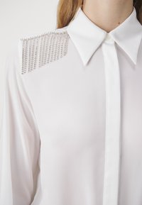 Blusa bianca con colletto appuntito e dettagli decorativi in perline d'argento sulle spalle. Tessuto liscio con chiusura a bottone sul davanti.