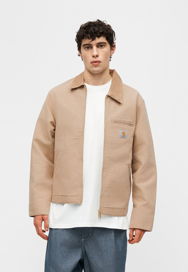 Giovane uomo con capelli ricci scuri che indossa una giacca beige con cerniera sopra una T-shirt bianca e jeans blu larghi, in piedi contro uno sfondo neutro.