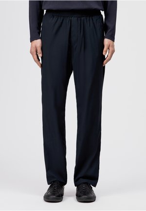 Mann trägt schwarze Freizeithose und schwarze Schnürschuhe, steht vor hellem einfarbigem Hintergrund.