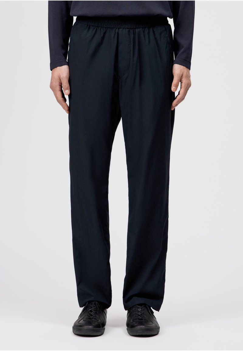 Homme portant un pantalon décontracté noir et des chaussures à lacets noires, debout devant un fond clair uni.