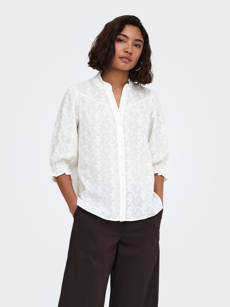 Mujer con cabello hasta los hombros que lleva una blusa blanca con estampado y mangas tres cuartos, y pantalones negros de pierna ancha, con las manos en los bolsillos, de pie.