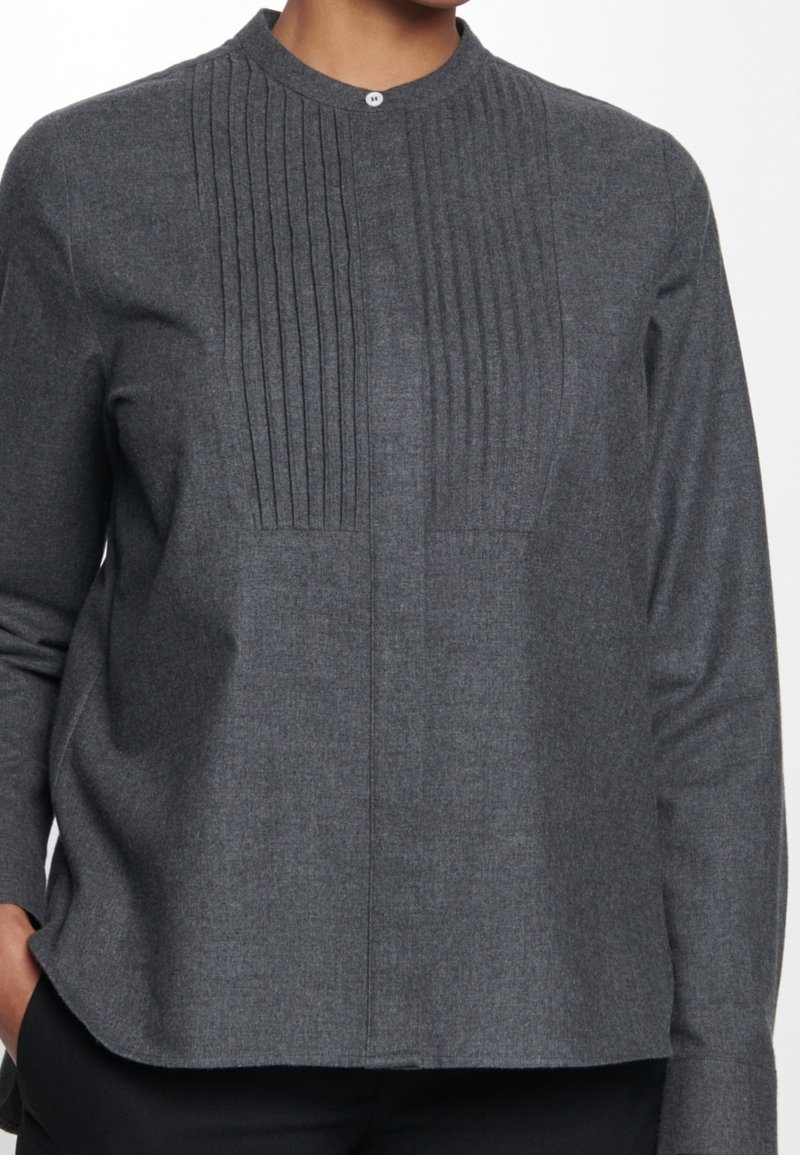 Personne portant une chemise gris foncé à manches longues avec des plis verticaux sur le devant et un seul bouton au col, main dans la poche.