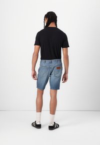 Vīriešu zilie denim šorti ar taisnu griezumu, redzamu šuvēm un brūnu ādas plāksteri uz aizmugurējā jostasvietas. Melna T-kreklu un sporta apavi.