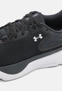 Chaussure de sport noire avec une tige en mesh, des accents gris texturés et une semelle blanche. Présente un laçage et le logo Under Armour sur le côté.