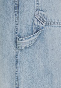 Ljusblå denimtyg med detaljerad sömnad och en liten, vikt bältesöglan nära sömmen på en bakficka.