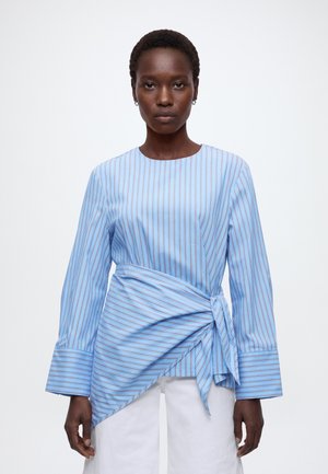 Woman wearing a long-sleeve blue striped blouse with a side tie detail and white pants, standing against a plain background.