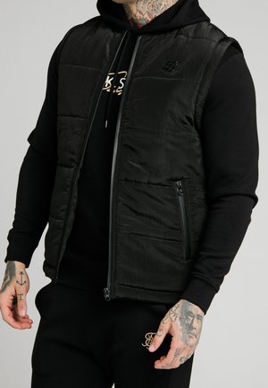 Uomo che indossa un gilet trapuntato nero senza maniche sopra una felpa nera con cappuccio, mostrando mani e collo tatuati, con dettagli di cerniera e logo sull'abbigliamento.