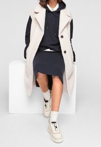 Cappotto senza maniche beige chiaro sopra un felpa blu navy e una gonna, abbinato a sneakers crema. Tessuto strutturato con bottoni neri e tasche laterali.