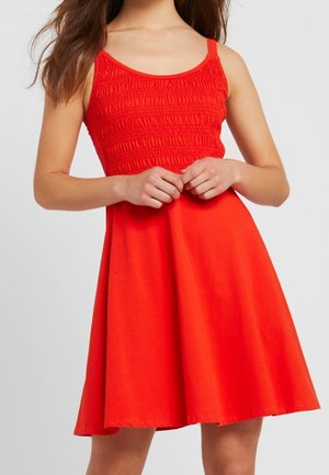 Młoda kobieta w bezrękawną, jaskrawoczerwoną sukienkę z teksturowanym gorsetem i rozkloszowanym dołem, ręce swobodnie złączone na talii.