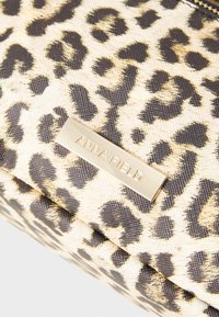 Kosmetiktasche mit Leopardenmuster aus strukturiertem Stoff, mit einem rechteckigen Logo-Plättchen in Goldtönen, auf dem "ANNA FIELD" steht. Sie zeigt ein Muster in Schwarz und Creme.