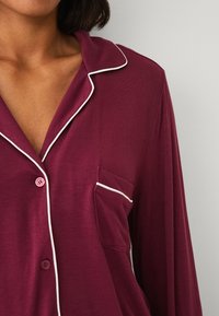 Burgundská pyžamová blůza s dlouhým rukávem a bílými lemy kolem límce a kapsy, s knoflíkovým zapínáním a hladkou texturou látky.