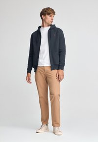 Ciemnoszara bluza z kapturem na zamek z troczkami, noszona na białej koszulce, w połączeniu z jasnobrązowymi spodniami i kremowymi trampkami.