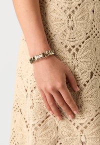 Gullfarget metallarmbånd med sammenkoblede kjedeledd, med en rektangulær lås. Hånd som hviler på et heklet beige stoff.