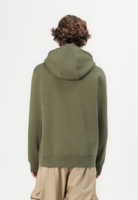 Hoodie verde-oliva com um grande capuz arredondado, textura suave, punhos e bainha canelados, e um corte descontraído, usado sobre calças cargo bege.
