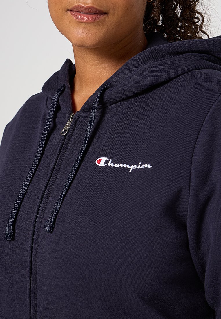 Felpa con zip di colore blu navy con cordini, presente un logo Champion ricamato in bianco sul lato sinistro del petto. Tessuto in morbido cotone misto.