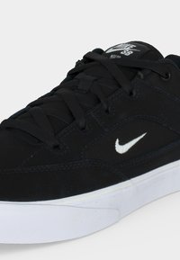 Μαύρο παπούτσι Nike SB με λευκό λογότυπο swoosh στο πλάι, μαύρα κορδόνια και λευκή σόλα, εμφανιζόμενο σε κοντινή γωνία.