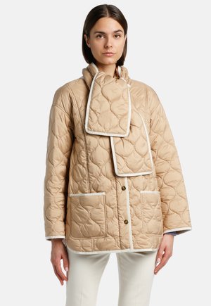 Beige gevoerde jas met een hoge kraag, witte piping, twee voorzakken en een unieke sjaalvormige detail bij de nek.