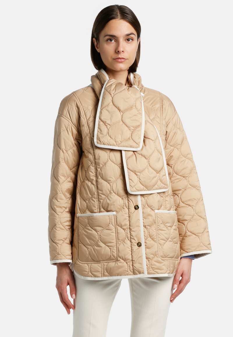 Giacca trapuntata beige con colletto alto, finiture bianche, due tasche frontali e un dettaglio unico a forma di sciarpa sul collo.
