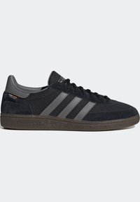 Svarta Adidas-sneakers med grå tre ränder, överdel av mocka och mesh, brunt texturerat sula och Cordura-typklistermärke på hälen.