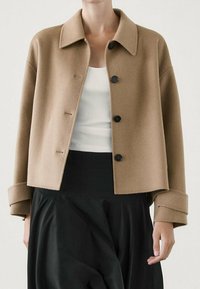 Beige wool-blend cropped jacket with a pointed collar, four black buttons, and wide sleeves, worn over a white tank and black skirt.