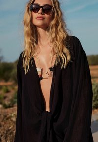 Ľahký čierny kimono štýl s viazaním vpredu, nosený cez viacfarebný bikini, doplnený slnečnými okuliarmi s korytnačkovým vzorom.