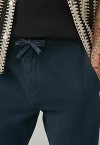 Pantaloni relaxați, de culoare albastru marin, cu talie elastică, având o buzunar frontal și un șnur legat. Materialul mai texturat este vizibil.