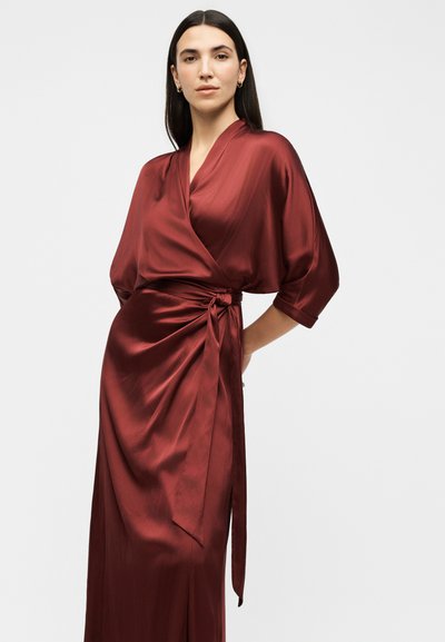 Femme portant une longue robe portefeuille bordeaux en soie avec des manches arrivant aux coudes, nouée à la taille, debout devant un fond clair uni.