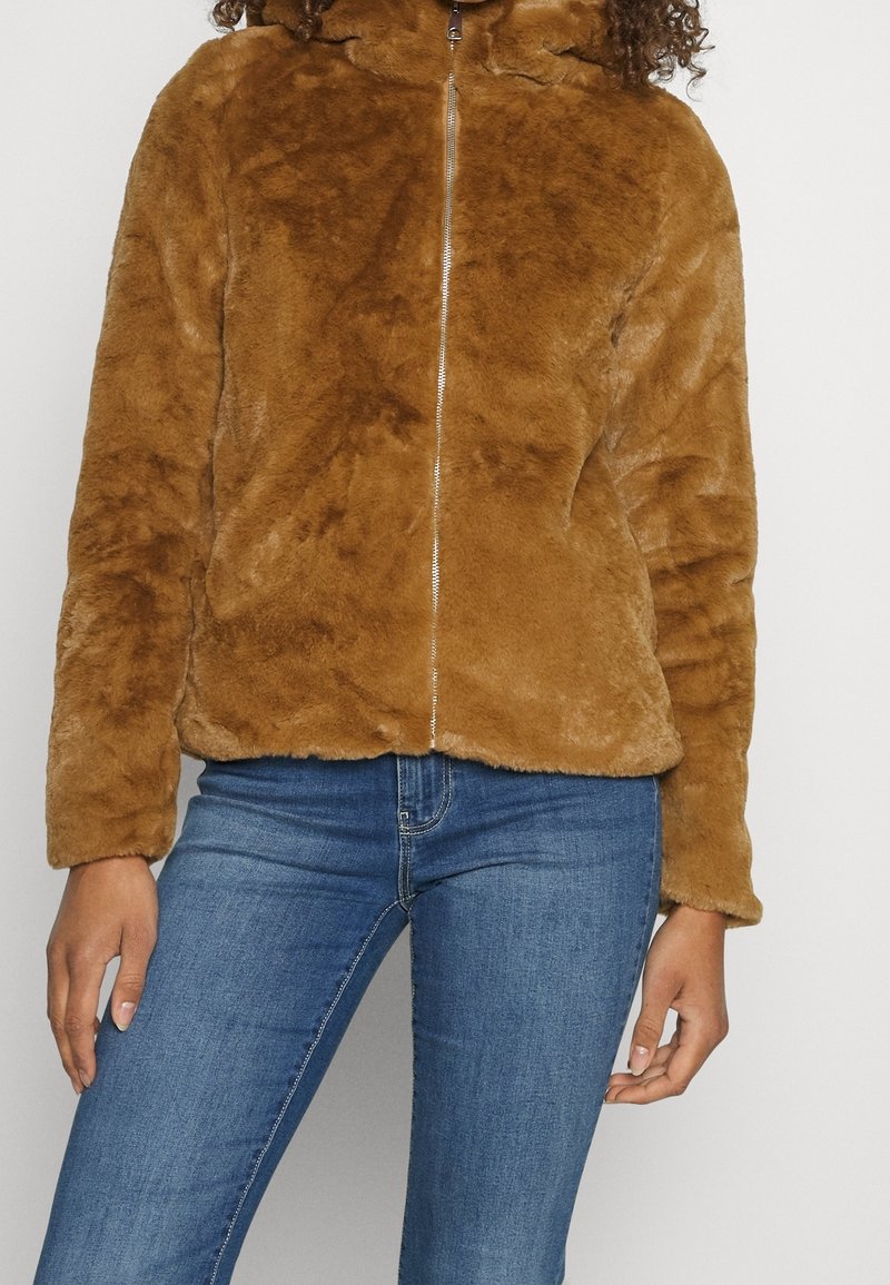 Personne portant une veste zippée en fausse fourrure marron et un jean bleu, debout devant un fond clair uni.