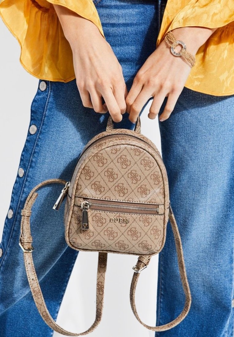 Mains tenant un petit sac à dos beige à motif Guess avec des bretelles réglables, porté avec un jean bleu et un haut jaune à manches longues.