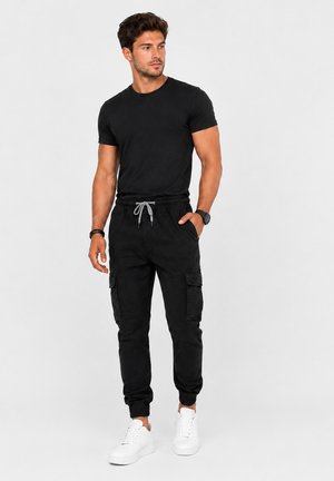 Mann mit dunklen Haaren, der ein eng anliegendes schwarzes T-Shirt, schwarze Cargo-Jogginghosen mit grauem Kordelzug, weiße Sneaker und eine schwarze Armbanduhr trägt.