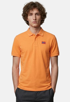 Junger Mann mit lockigem Haar, der ein orangefarbenes Poloshirt mit einem kleinen norwegischen Flaggenabzeichen auf der Brust und dunkelgraue Hosen trägt.