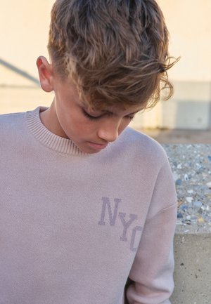 Chlapec so svetlohnedými vlasmi sa díva dolu, má na sebe svetloružový sveter s potlačou "NYC" na hrudi, stojí vedľa betónovej steny vonku.