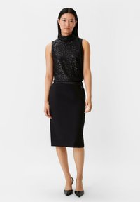 Schwarzes ärmelloses Oberteil mit Pailletten-Design, kombiniert mit einem taillierten schwarzen Bleistiftrock. Das Model trägt schwarze spitze Pumps.