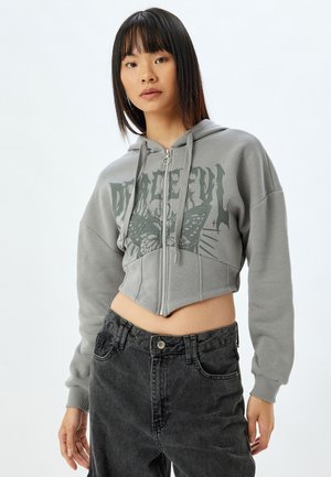 Geknipte grijze hoodie met een vlinderafbeelding, ritssluiting en een capuchon met trekkoord. Gecombineerd met hoge zwarte jeans.