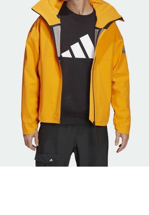 Homme portant une veste jaune vif dézippée sur un t-shirt noir avec un logo Adidas blanc audacieux, associé à un pantalon noir avec une poche latérale.