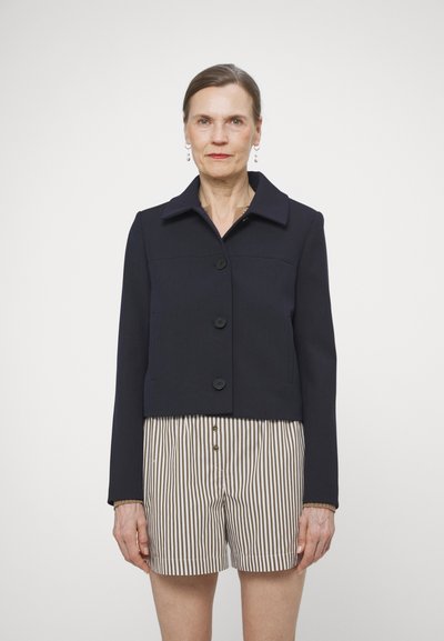 Veste cropped navy avec un col pointu, comportant quatre boutons et une finition soignée. Le mannequin porte des shorts rayés en dessous.