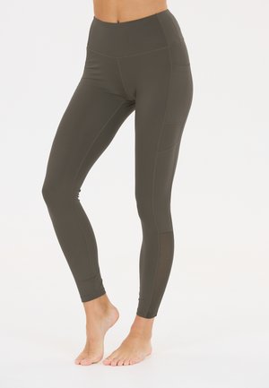 Frau trägt hoch taillierte dunkelgraue Leggings mit Seitentaschen und steht barfuß vor einem schlichten weißen Hintergrund.