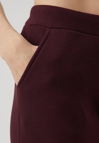 Burgunder Jogginghosen mit ribbeltem Bund und einer Seitentasche, aus einem weichen, texturierten Stoff. Nahaufnahme einer Hand in der Tasche.