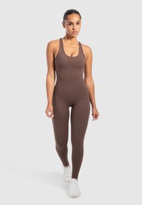 Vrouw die naar voren loopt in een aansluitende bruine mouwloze jumpsuit en witte sportschoenen tegen een effen grijze achtergrond.