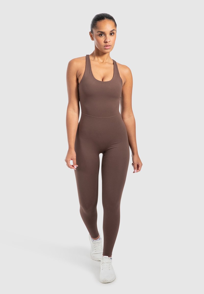 Vrouw die naar voren loopt in een aansluitende bruine mouwloze jumpsuit en witte sportschoenen tegen een effen grijze achtergrond.