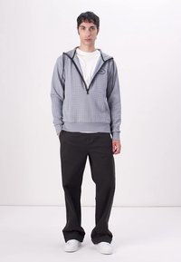 Sudadera gris a cuadros con cremallera en la parte delantera, ribetes negros en contraste y un logo. Combinada con pantalones anchos negros y zapatillas blancas.