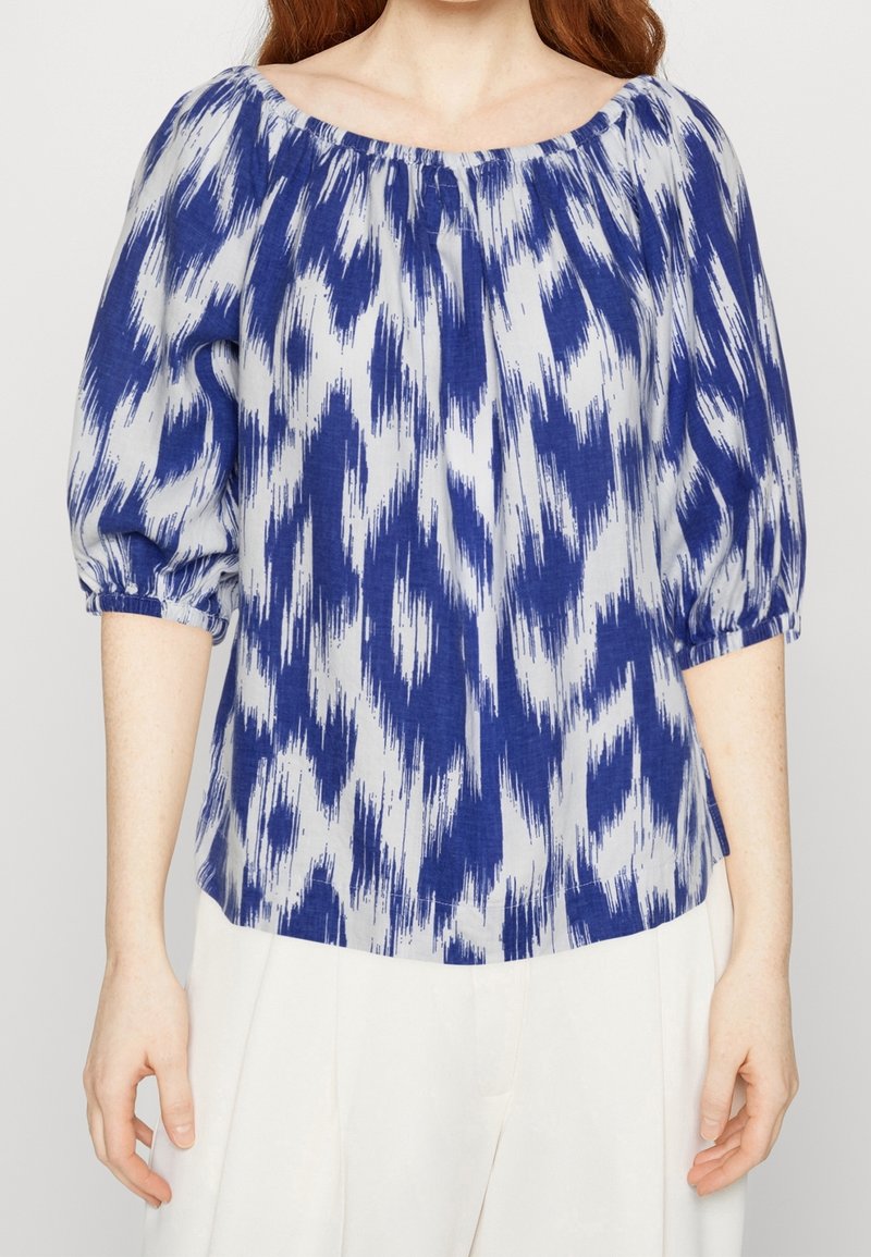 Blusa blu e bianca con scollo off-the-shoulder, maniche a sbuffo e motivo ikat. Tessuto leggero con una vestibilità rilassata.