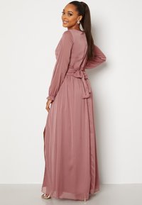 Dlouhé růžové šaty s dlouhými rukávy, vázaným pasem a volnou sukní s bočním rozparkem. Hladká, lehká látka s jemným leskem.